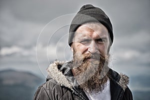 Man with long beard and mustache wears hat. Hipster on strict face with beard looks brutally while hiking. Masculinity