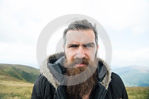 Man with long beard and mustache wears jacket. Hipster on strict face with beard looks brutally while hiking. Hermit