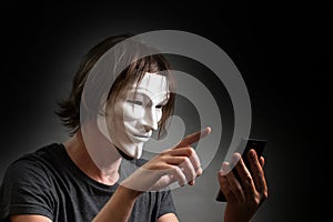 A man with long hair in a hacker or anonymous mask in a T-shirt is holding a mobile phone in his hands. The concept of an Internet