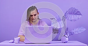 A man with long hair is sitting at a computer in a lilac-colored room and drinking a drink. Creative space for