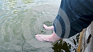 Man in long shorts dangles his legs in the water of lake. splashing water