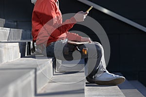 Man longboarder in casual clothes using his smartphone, resting on the steps, sitting with longboard/skateboard outdoors