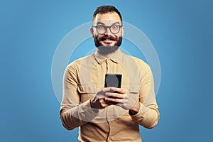 Man looking at camera while holding mobile phone, isolated over blue background