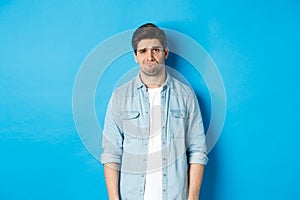 Man looking with doubt at camera, frowning and sulking, feeling uneasy about something, standing against blue background