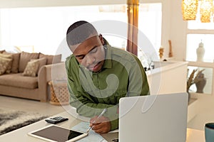 Man looking at laptop screen and writing on paper at dining table in kitchen at comfortable home