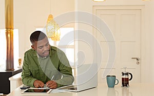 Man looking at laptop screen and writing on paper at dining table in kitchen at comfortable home