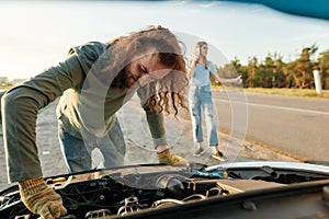 Man looking under the hood of broken car, girl calling car service or assistance in the background