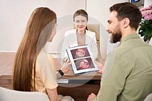 Man looking at wife looking at ultrasound scan of her pregnant belly near doctor
