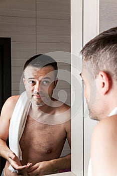 A man looks in the bathroom mirror in the morning with a towel on his shoulder