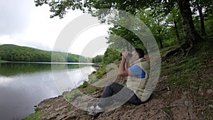 Man looks through binoculars at distance while sitting at scenic river bank on green grass in shadow of tree.