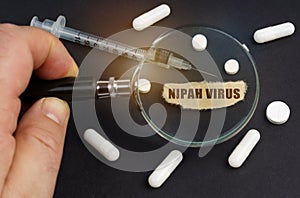 A man looks through a magnifying glass at pills, a syringe and a piece of paper with the inscription - Nipah virus