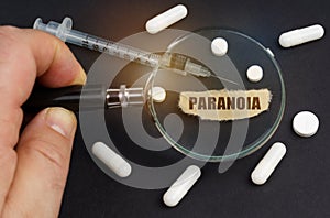 A man looks through a magnifying glass at pills, a syringe and a piece of paper with the inscription - paranoia