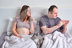 A man looks at the phone on the bed next to his pregnant wife, relationship problems during pregnancy