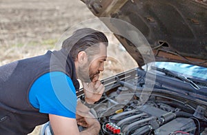 A man looks thoughtfully at the engine under the hood during a breakdown on a rural road.
