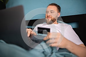 Man lying in bed doing online shopping using credit card and laptop while relaxing in bed on weekend