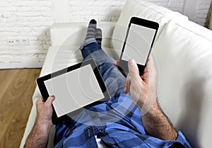 Man lying on couch at home living room using simultaneously mobile phone and digital tablet pad in internet technology