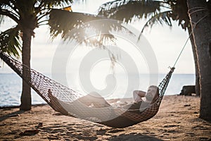 A man is lying in a hammock at sunset by the ocean. Handsome guy is resting in a hammock among palm trees on the seashore
