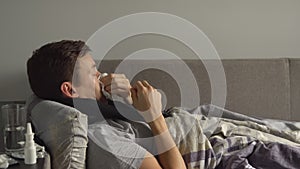 Man lying sick in bed next to his medications in his home and blowing his nose in a handkerchief