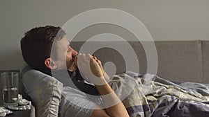 Man lying sick in bed next to his medications in his home and blowing his nose in a handkerchief