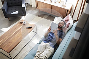 Man Lying On Sofa At Home Wearing Headphones And Watching Movie On Digital Tablet