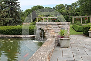 A man made stone waterfall flowing into a large koi pond with stone pavers leading to a landscaped garden