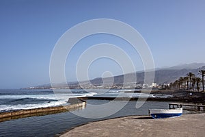 A man-made swimming pool nearby the ocean, a small fishing boat and a  boardwalk leading towards the resorts of Costa Adeje