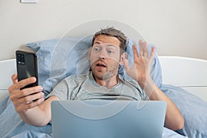 Man making video call on smartlphone waving hello communicating online while lying in bed with laptop in a bed at home