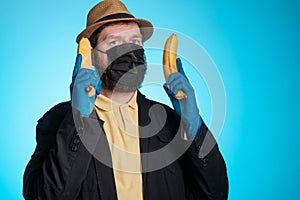 A man in a mask and a hat holds bananas in both hands like a weapon, the background is blue