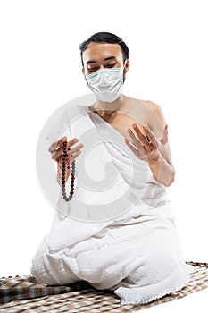 Man in mask wearing ihram clothes praying with prayer beads