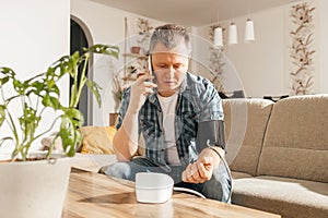 A man measures blood pressure at home with a blood pressure cuff and a tonometer. A man calls a doctor on a mobile phone