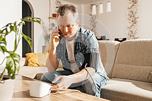 A man measures blood pressure at home with a blood pressure cuff and a tonometer. A man calls a doctor on a mobile phone