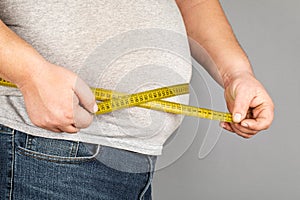 A man measures his fat belly with a measuring tape. on a gray background