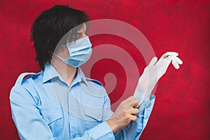 Man in a medical mask puts on protective gloves on a red background. male portrait. virus protection measures