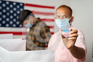 Man in medical mask showing I voted Sticker at polling booth with US flag as background - concept in person voting at US election