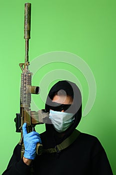 Man in a medical mask and sunglasses dressed in black clothes with a hood holds an automatic assault weapon