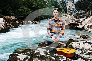 Man in a meditative position with guitar sitting on the bank of a mountain river on a background of rocks and forest. Concept of