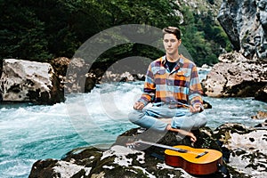 Man in a meditative position with guitar sitting on the bank of a mountain river on a background of rocks and forest. Concept of