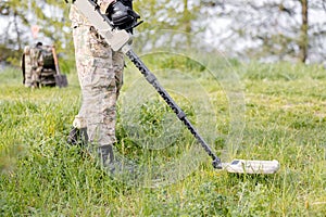 A man in a military uniform and bulletproof vest works in the forest with a metal detector. A minesweeper performs work on