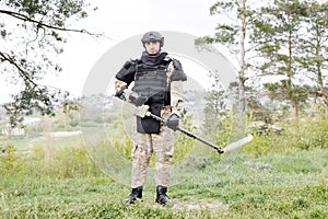 A man in a military uniform and bulletproof vest works in the forest with a metal detector. A minesweeper performs work on