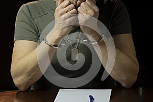 A man in military uniform with handcuffs on his wrists sits at a desk, pondering a legal document,