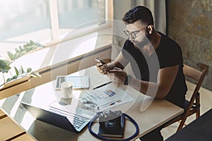 Man millenial in glasses sits at table in front of laptop and uses smartphone, reads message from business partner