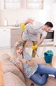 Man mopping floor while woman resting on sofa