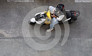 Man on motor scooter wear hardhat, helmet, delivering fast food in yellow thermal box. guy looking in mobile phone. top aerial