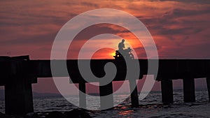 A man on a motorcycle taking selfies on a bridge over the ocean at sunset.