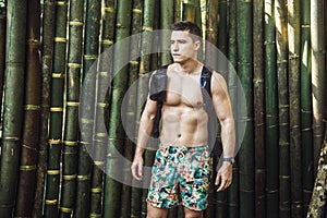 Man with muscle body wearing swimming shorts and backpack at tropical bamboo background