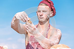 Man muscular baker or cook covered with flour working outdoor, sky on background. Baker adds egg to prepare dough