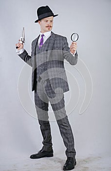 Man with a mustache dressed in a business plaid suit with a tie, holding a magnifying glass and a gun on a white solid Studio back