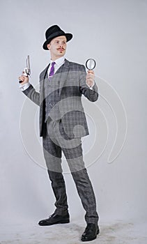 Man with a mustache dressed in a business plaid suit with a tie, holding a magnifying glass and a gun on a white solid Studio back
