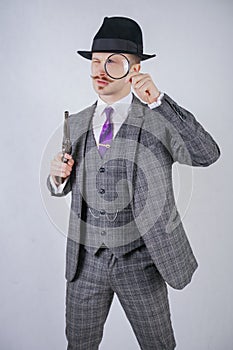 Man with a mustache dressed in a business plaid suit with a tie, holding a magnifying glass and a gun on a white solid Studio back