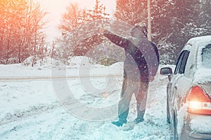 Man near broken car on winter road rises hand asking for help, breakdown of transport on country road in snow blizzard, copy space